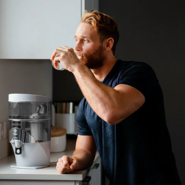 Man enjoying a glass of filtered water from the NIKKEN PiMag Waterfall Water Filtration System in a modern kitchen, showcasing premium hydration, cleaner drinking water, and everyday home wellness.